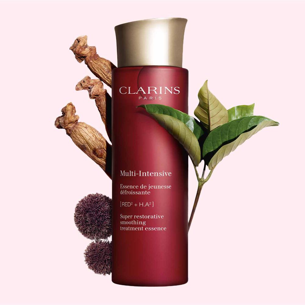 Packshot of Clarins Super Restorative Treatment Essence near red jania, Harungana leaves, and ginseng root on pink backdrop.