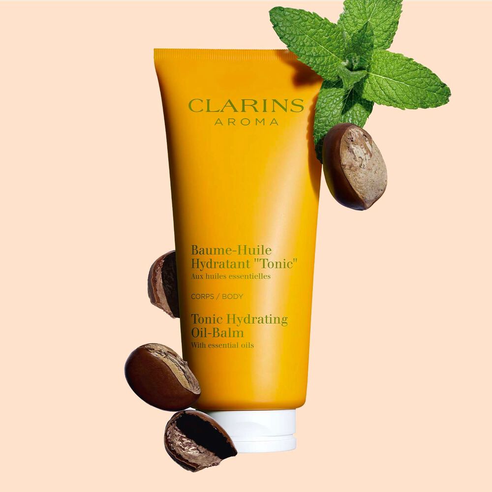 Packshot of a tube of Clarins Tonic Hydrating Oil-balm, close to some mint leaves and shea seeds on a peach background.