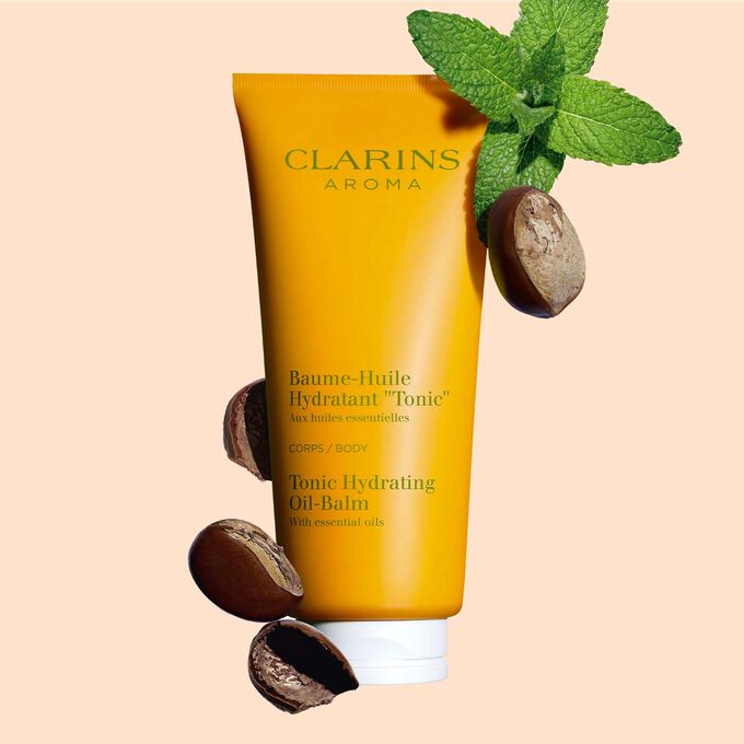 Packshot of a tube of Clarins Tonic Hydrating Oil-balm, close to some mint leaves and shea seeds on a peach background.