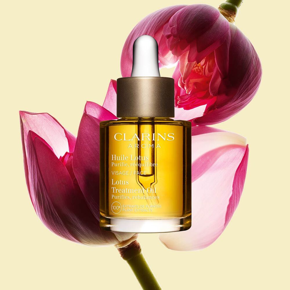 Packshot of a bottle of Clarins Lotus Face Treatment Oil close to a lotus flower on a pale yellow background.