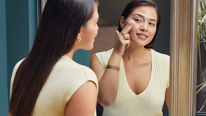Woman looking at herself in the mirror, wondering when to start using an anti-wrinkle cream to keep a  visible youthful skin